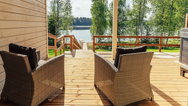 Una terraza de madera con muebles de mimbre y vistas a un lago y un bosque.