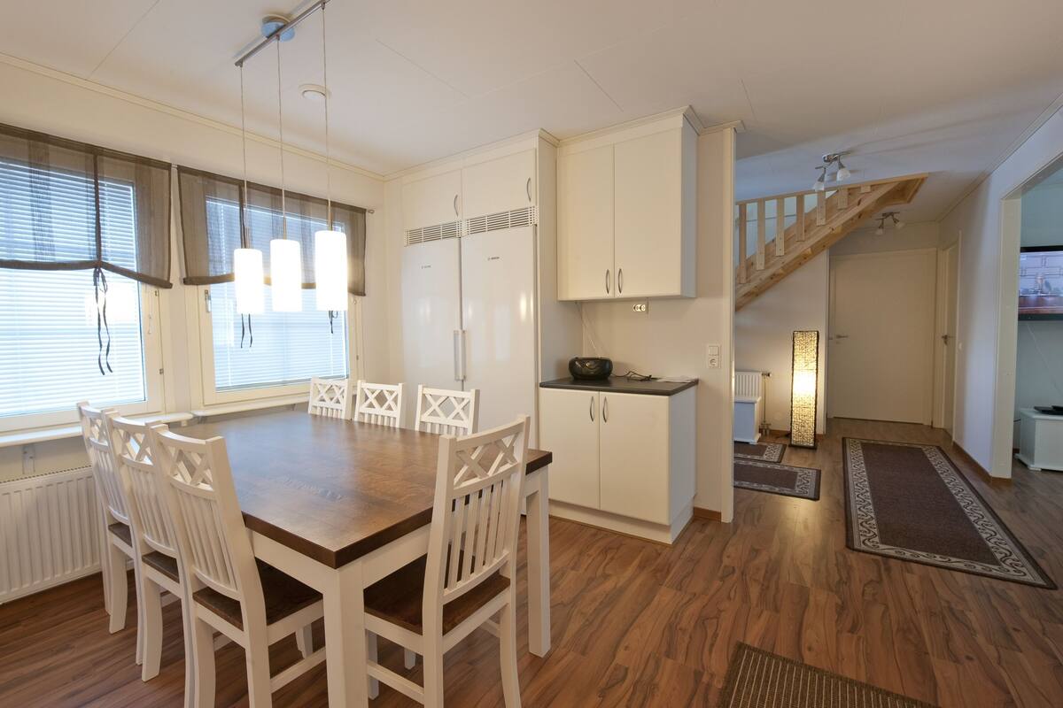 Eine moderne Küche mit weißen Schränken, einem Holzespeisetisch und weißen Stühlen. Eine Treppe führt zu einer weiteren Ebene.