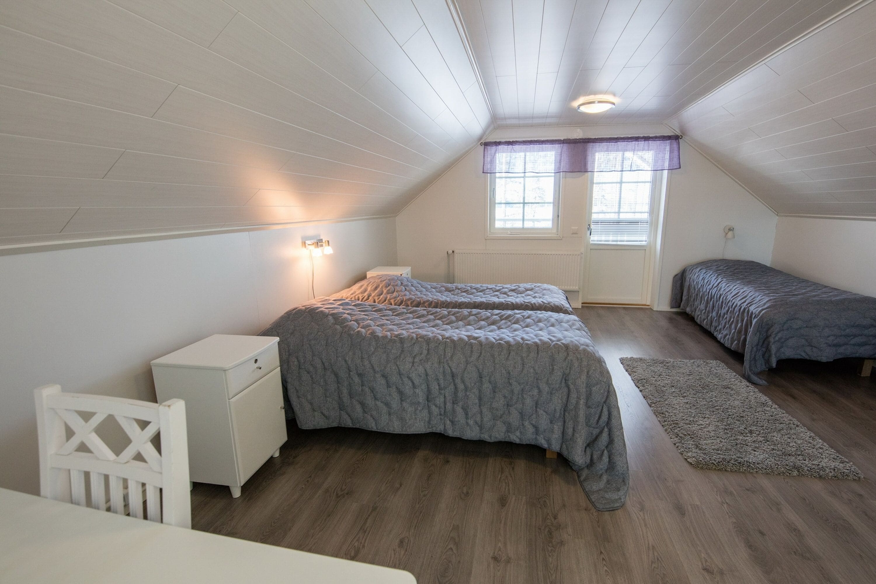 Un dormitorio con dos camas, una mesita de noche blanca, una silla blanca y una ventana con cortina púrpura.