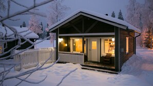 Una cabaña de madera con techo nevado, un porche con banca y un interior visible con comedor.