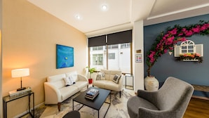 A modern living room with a beige sofa, a grey armchair, a coffee table, and a floor lamp. There is a painting on the wall, a potted plant, and a decorative wall piece with flowers. - Z Otel (Cesme)