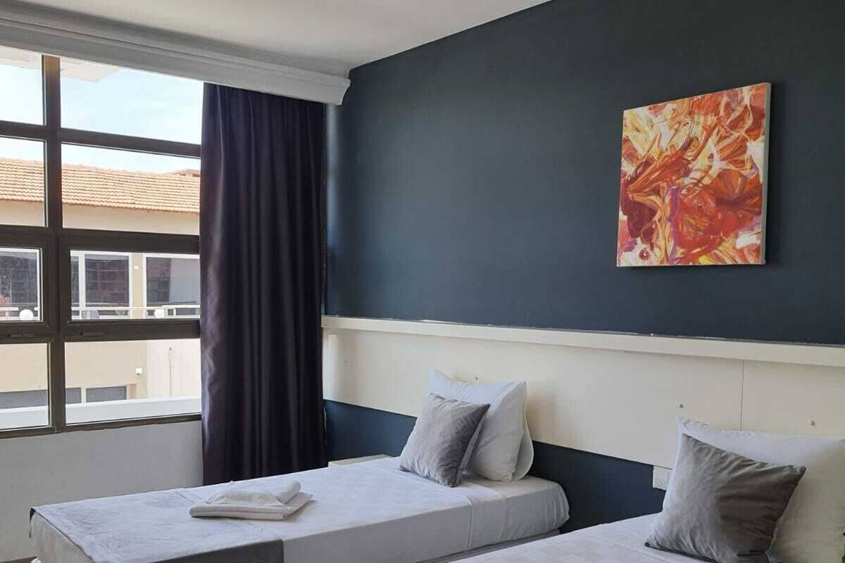 A hotel room with two single beds, a large window with curtains, a ceiling light, and a painting on the wall.