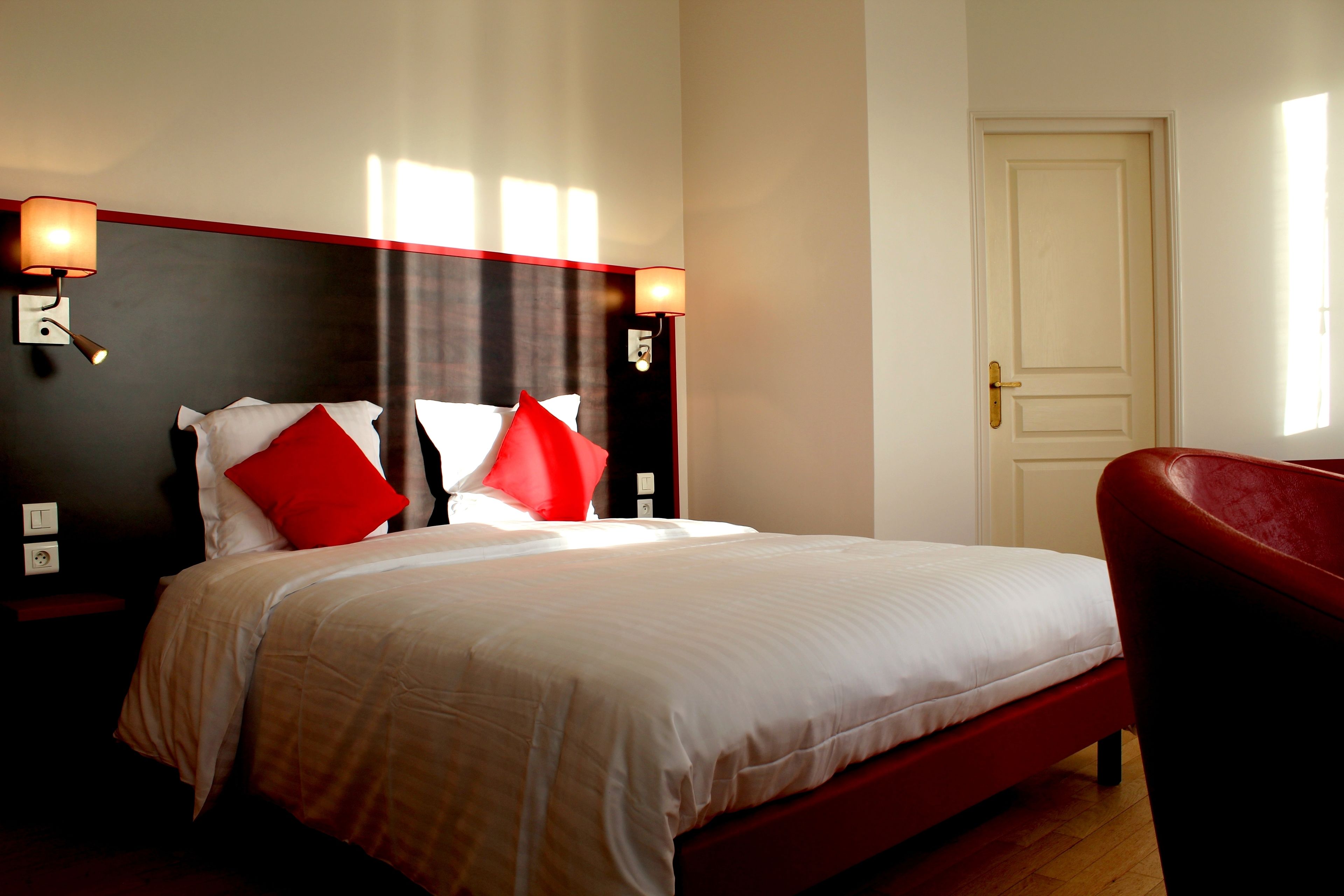Habitación de hotel con cama, dos mesitas de noche, un tocador, una silla roja y una puerta.