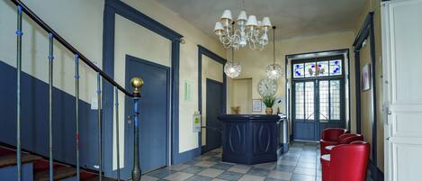 A hotel lobby with a checkered floor, a staircase with blue and white railings, a reception desk, and a chandelier.