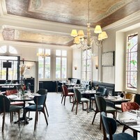 A restaurant with a classic interior, featuring a high ceiling with ornate plasterwork, large windows with stained glass, and a variety of seating arrangements including tables and booths.