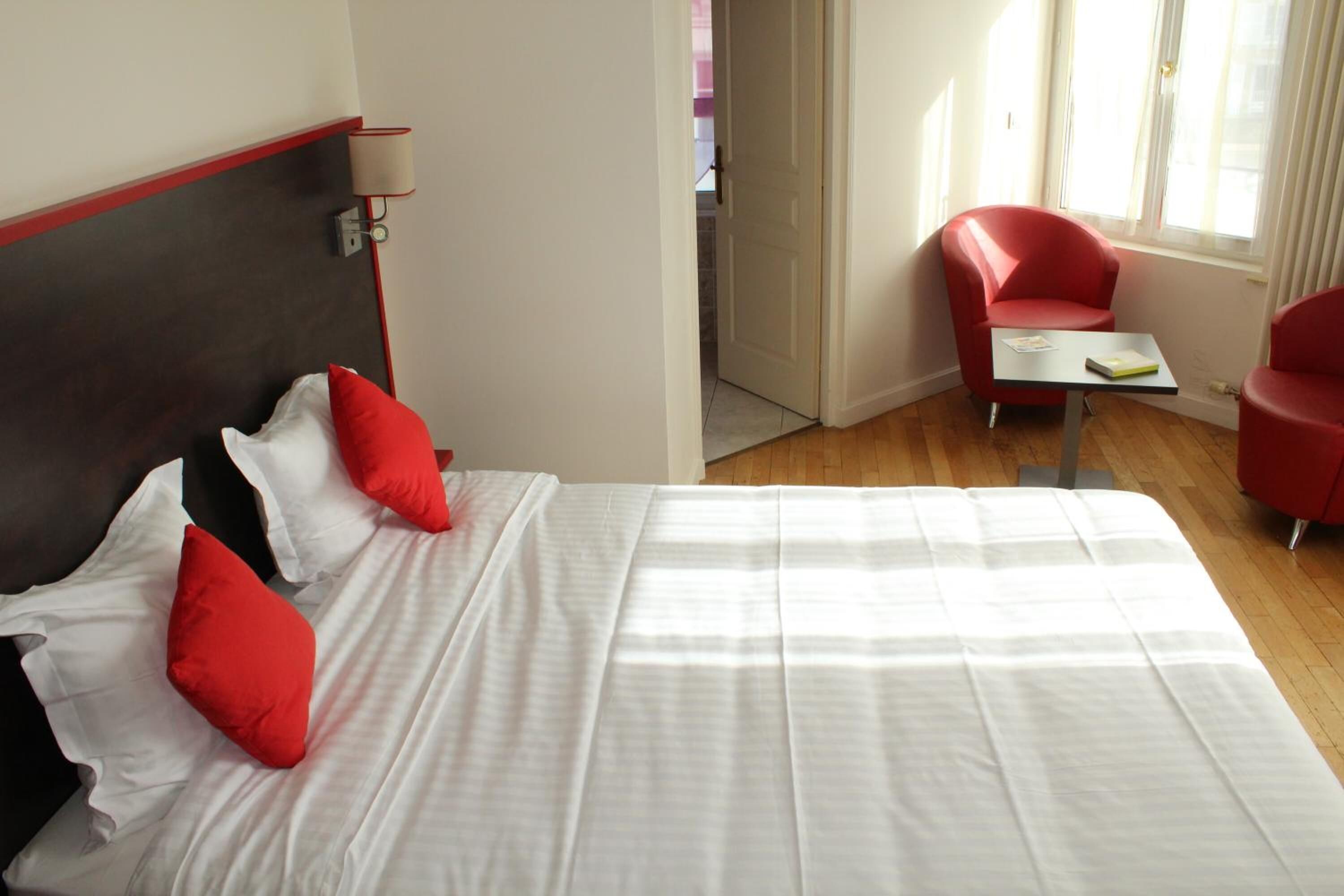 Habitación de hotel con cama, sillas rojas, una mesita y ventana con cortinas.