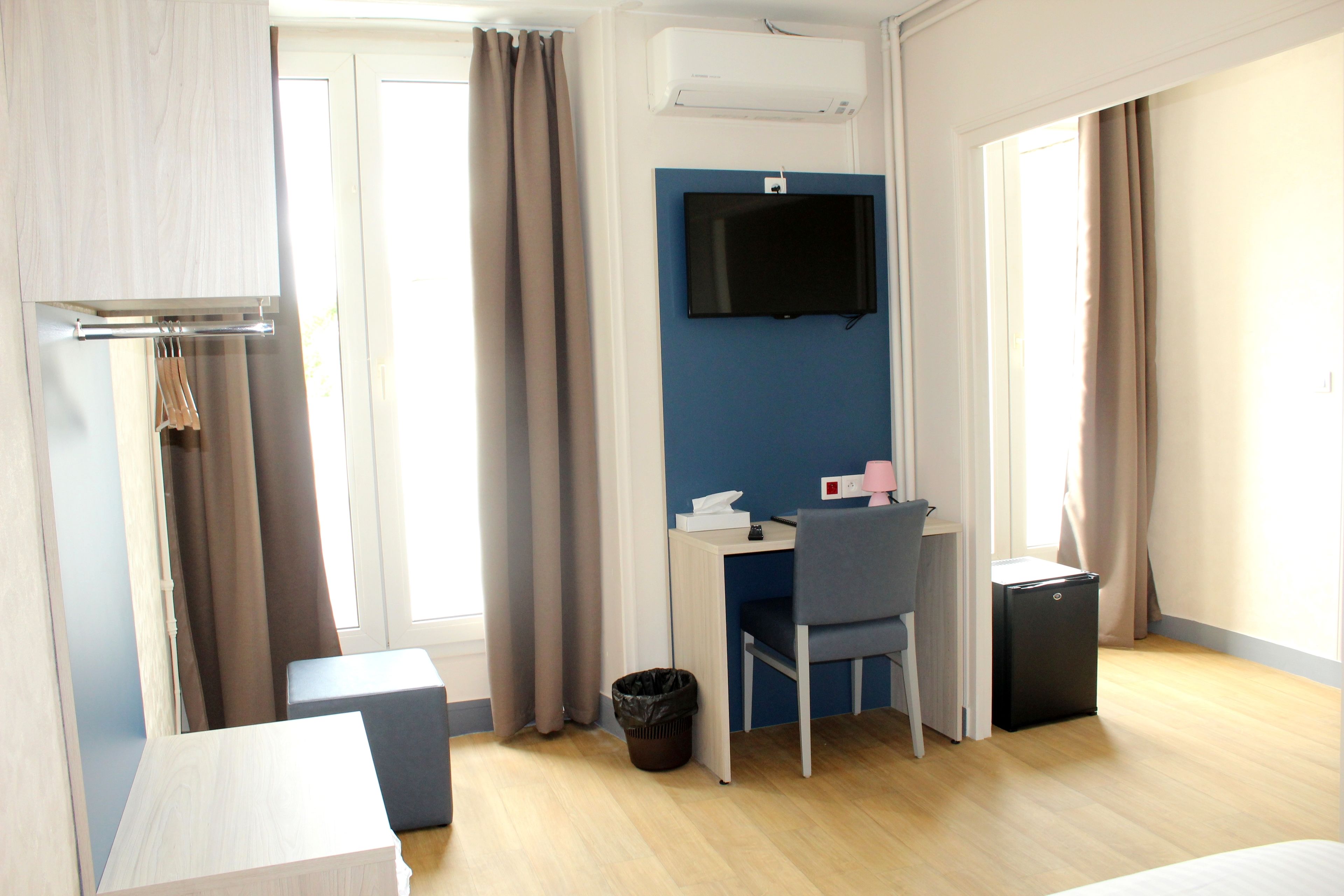 Une chambre d’hôtel moderne avec un plancher en bois, un bureau avec une chaise, un petit réfrigérateur noir, un téléviseur fixé au mur et une fenêtre avec des rideaux.