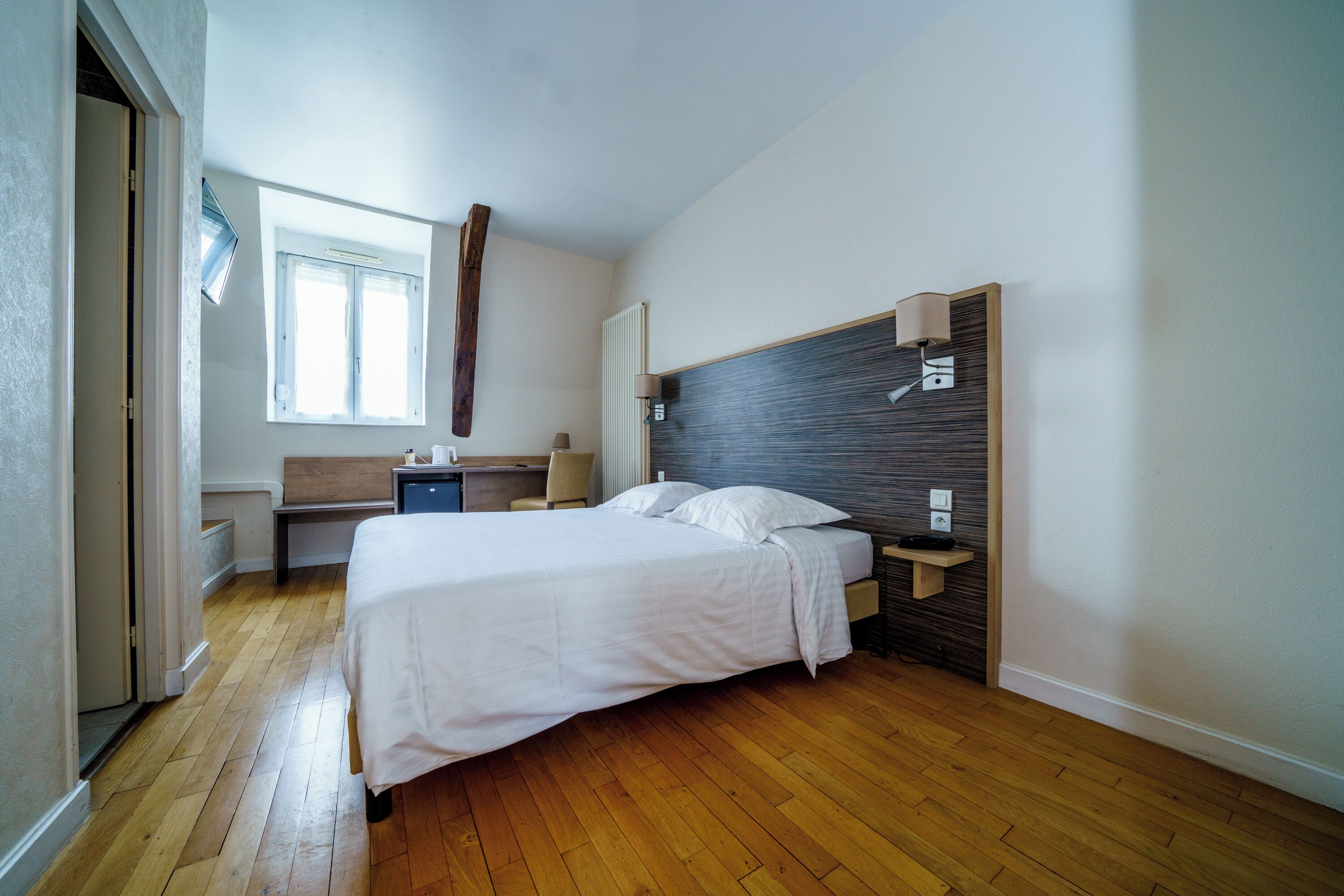 Ein Schlafzimmer mit Holzboden, einem Bett mit weißer Bettwäsche, einem Nachttisch, einem Schreibtisch, einem Stuhl, einem Fenster und einer Heizung.