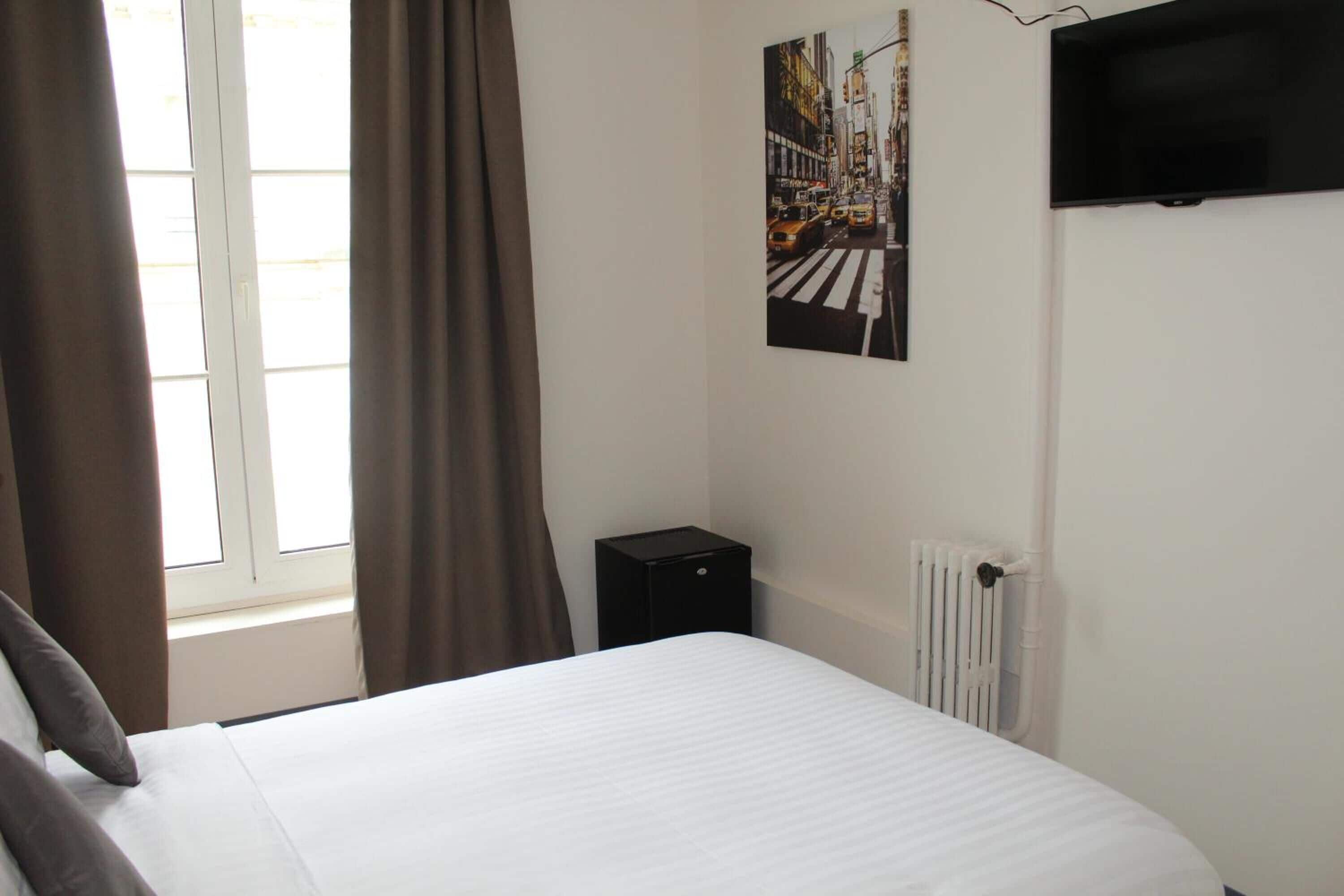 A hotel room with a bed, a radiator, a black speaker, and a cityscape painting on the wall.