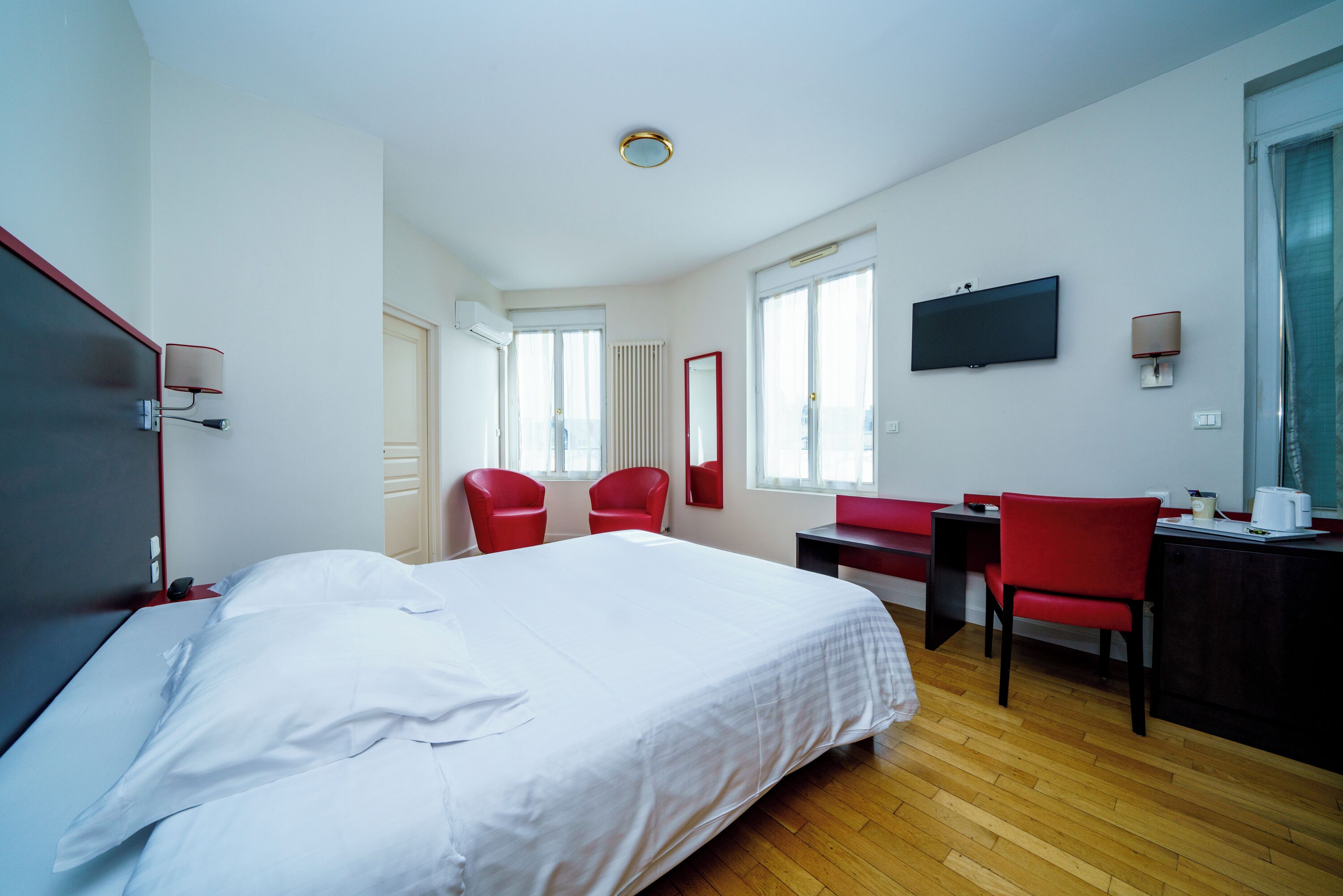 Una habitación de hotel con una cama, dos sillas rojas, un escritorio con una silla, un televisor y una ventana.