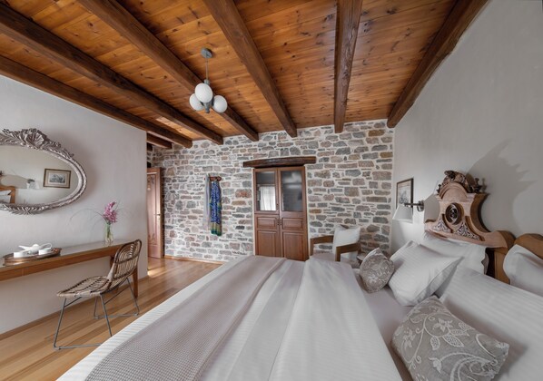 Un dormitorio con pared de piedra, vigas de madera en el techo, una cama con almohadas, una silla, un aparador y un espejo.