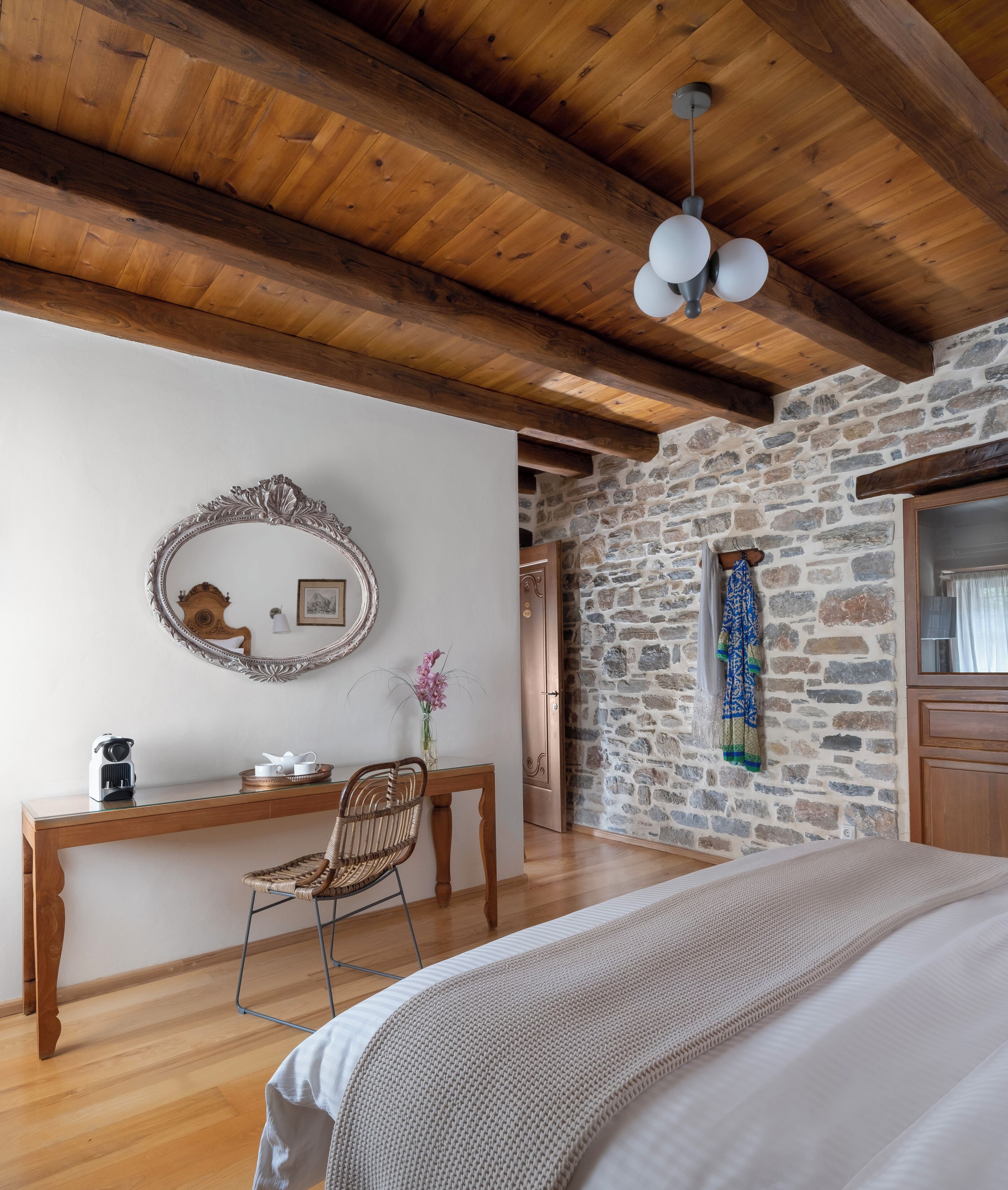 Une chambre avec un plafond en bois, un mur en pierre, un lit, un bureau, une chaise et un miroir.