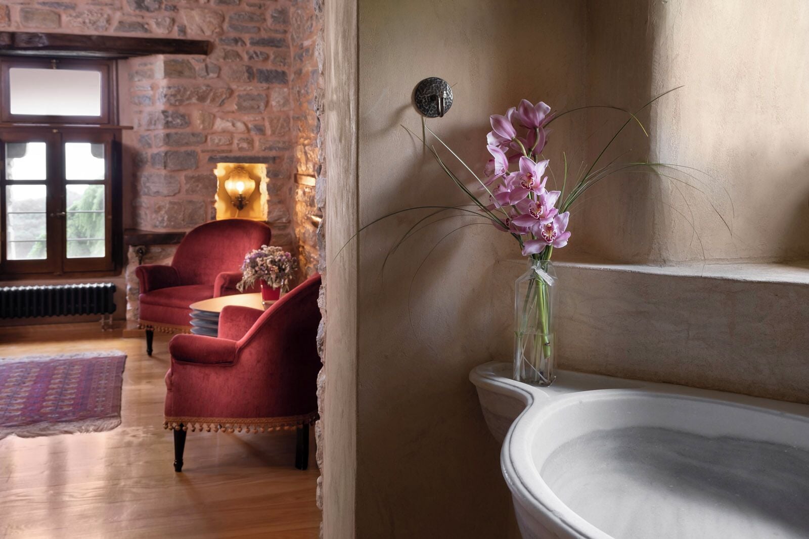 A room with a stone wall, a red armchair, a vase with pink flowers, and a white bathtub.