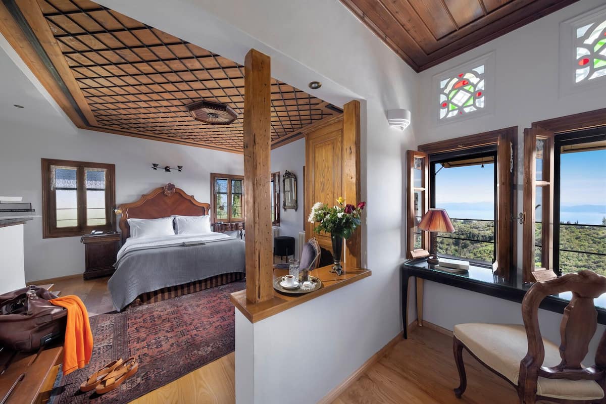 A bedroom with a wooden ceiling, a bed with a headboard, a nightstand, a desk with a lamp, a chair, and a view of the outdoors through large windows.
