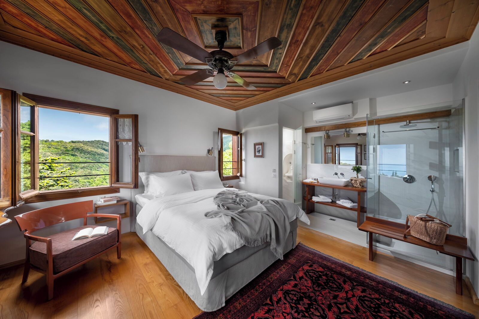 Ein Schlafzimmer mit einem großen Bett, Holzboden, einem Fenster mit Blick auf Grünflächen, einem Badezimmer mit Glasdusche und einem Nachttisch mit einem Buch.