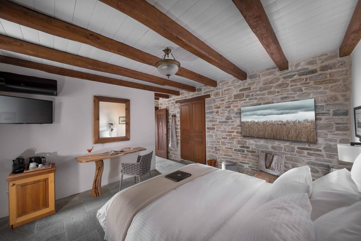 A bedroom with a stone wall, wooden beams, a bed, a desk, and a television.