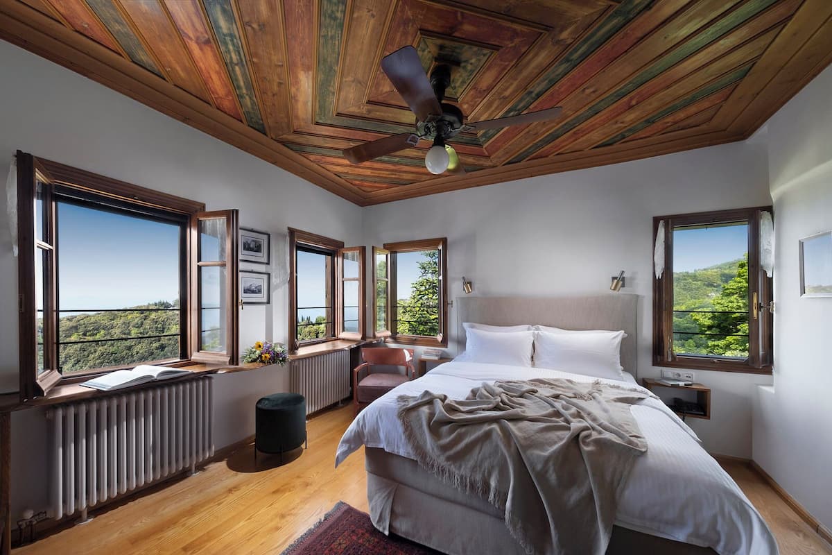 A bedroom with a large bed, multiple windows, a ceiling fan, and a view of a forested area.