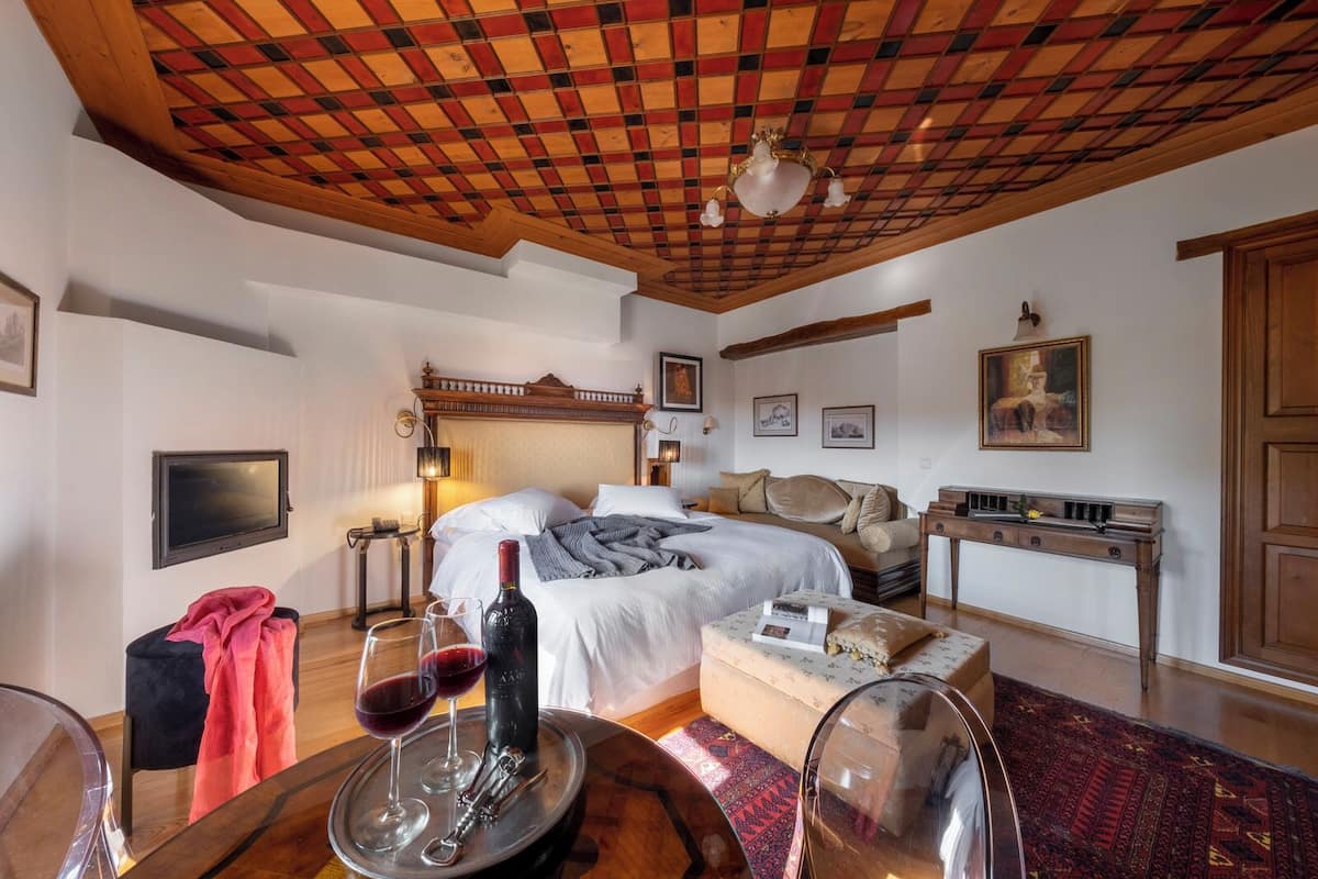 A bedroom with a wooden ceiling, a bed with white linens, a bedside table with a lamp, and a small table with wine glasses and a bottle.