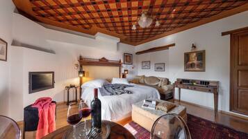 Quarto com teto de madeira, cama com roupa de cama branca, mesa de cabeceira com luminária e pequena mesa com copos de vinho e uma garrafa.