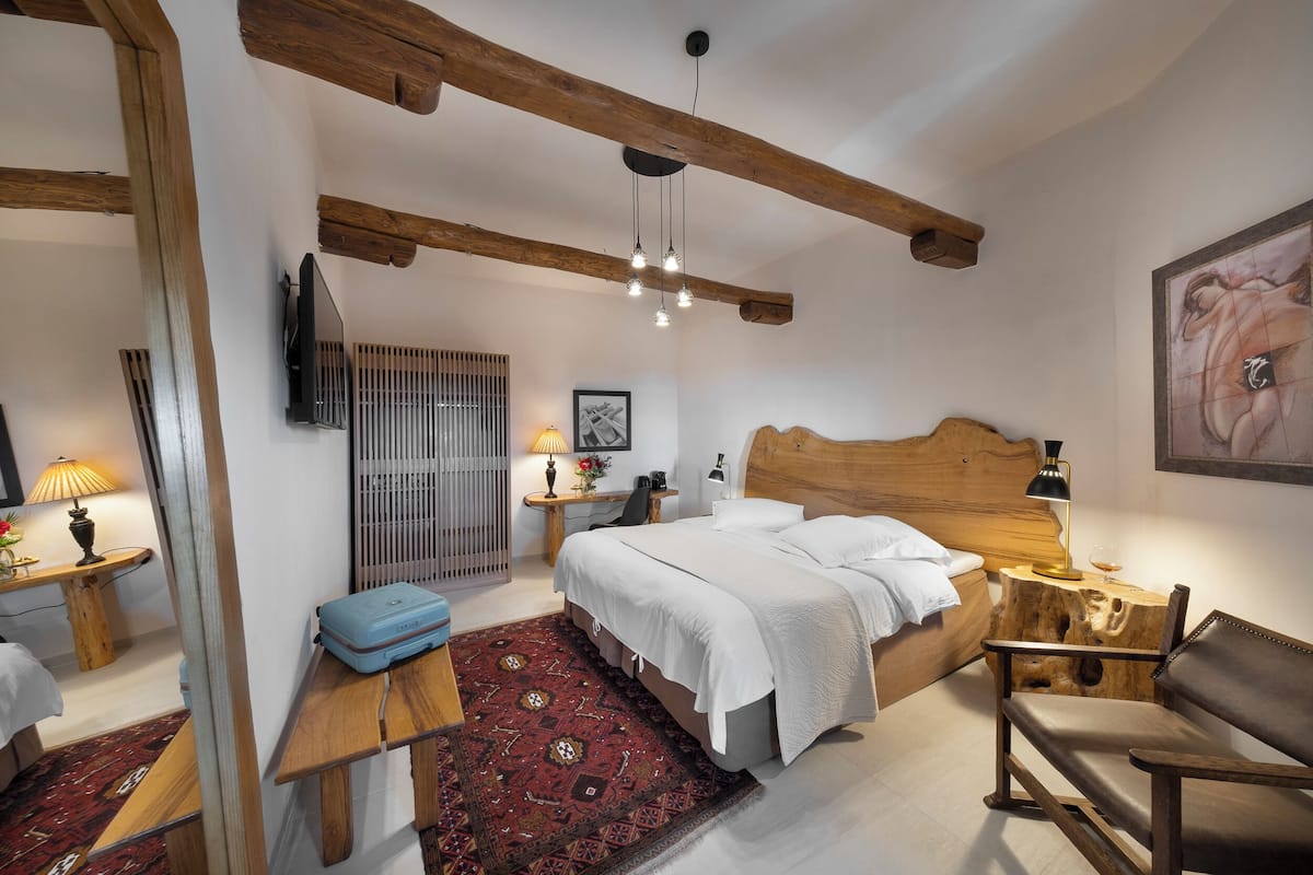 A bedroom with a wooden headboard, a bed with white linens, a wooden bench, a small table with a lamp, a red patterned rug, and a wooden floor.