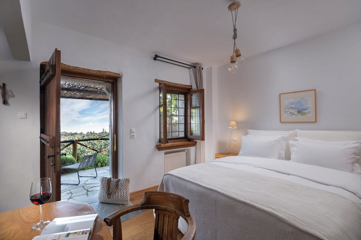 A bedroom with a bed, a wooden chair, a table with a glass of wine, and a view of the outdoors through an open door.