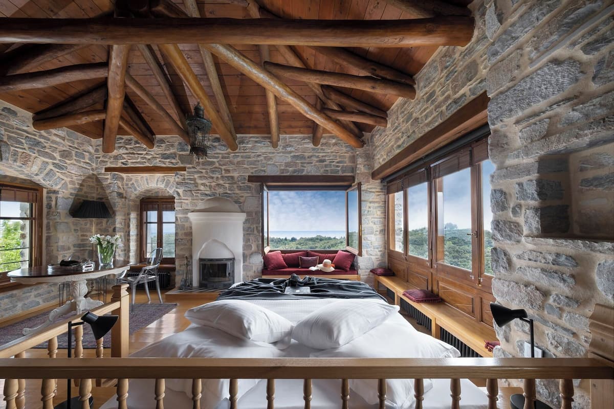 A rustic bedroom with a stone wall, wooden ceiling beams, a fireplace, a large bed, and a view of the landscape.