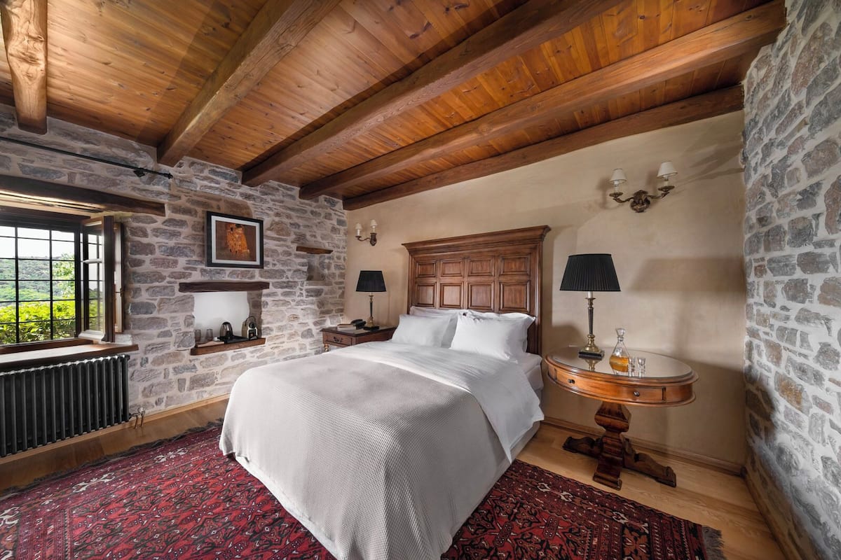 A cozy bedroom with a large bed, stone walls, wooden beams, a window with a view, and a nightstand with a lamp.