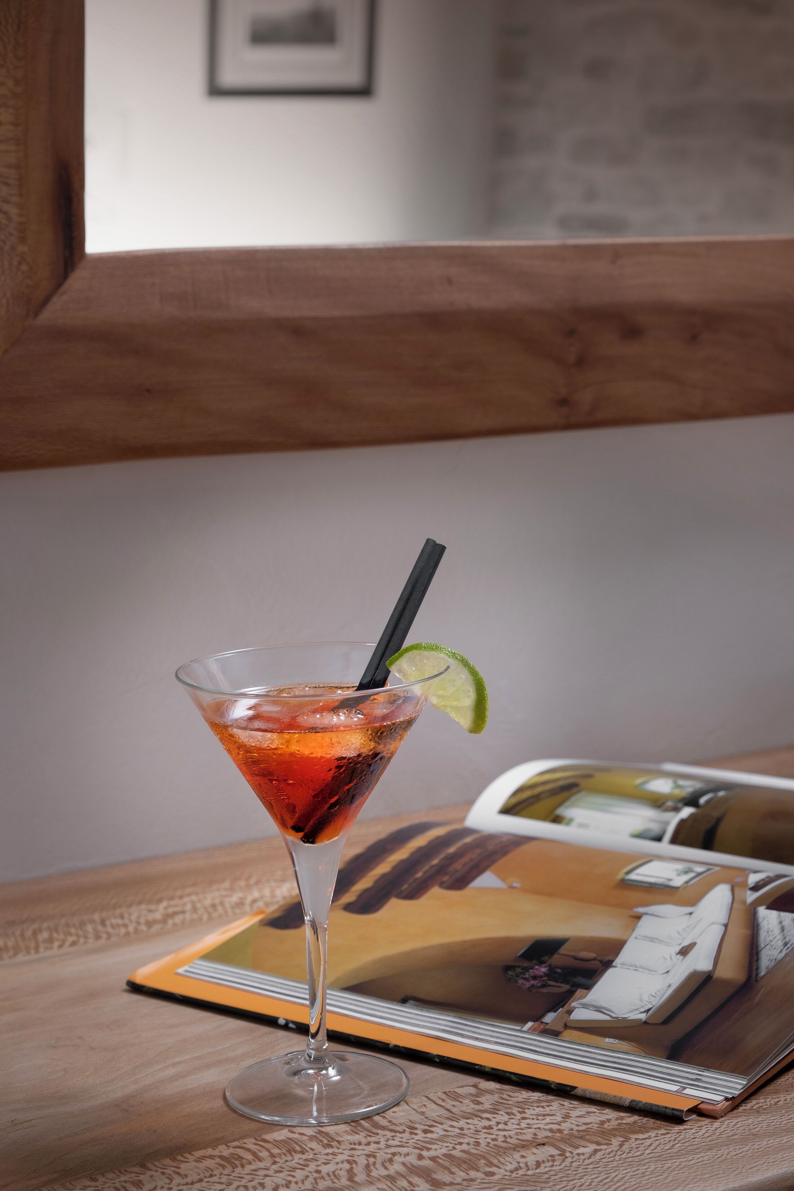 A martini glass with a lime wedge and black straw on a wooden table.