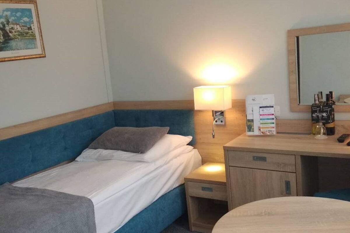 Ein Hotelzimmer mit einem Einzelbett, einem Nachttisch mit Lampe, einem kleinen Schreibtisch mit Spiegel und einem runden Tisch.