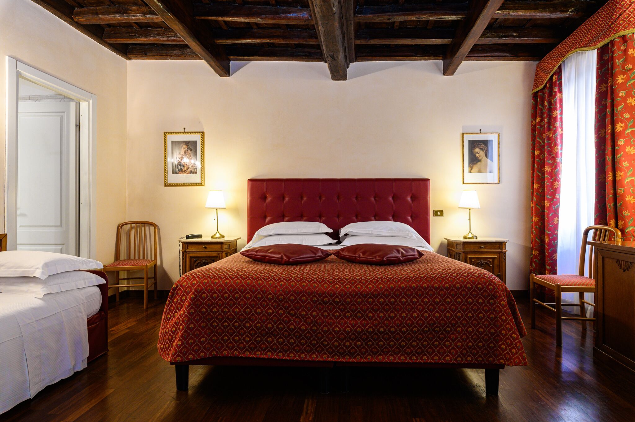 A bedroom with a red upholstered bed, two bedside tables with lamps, a wooden chair, and a door.