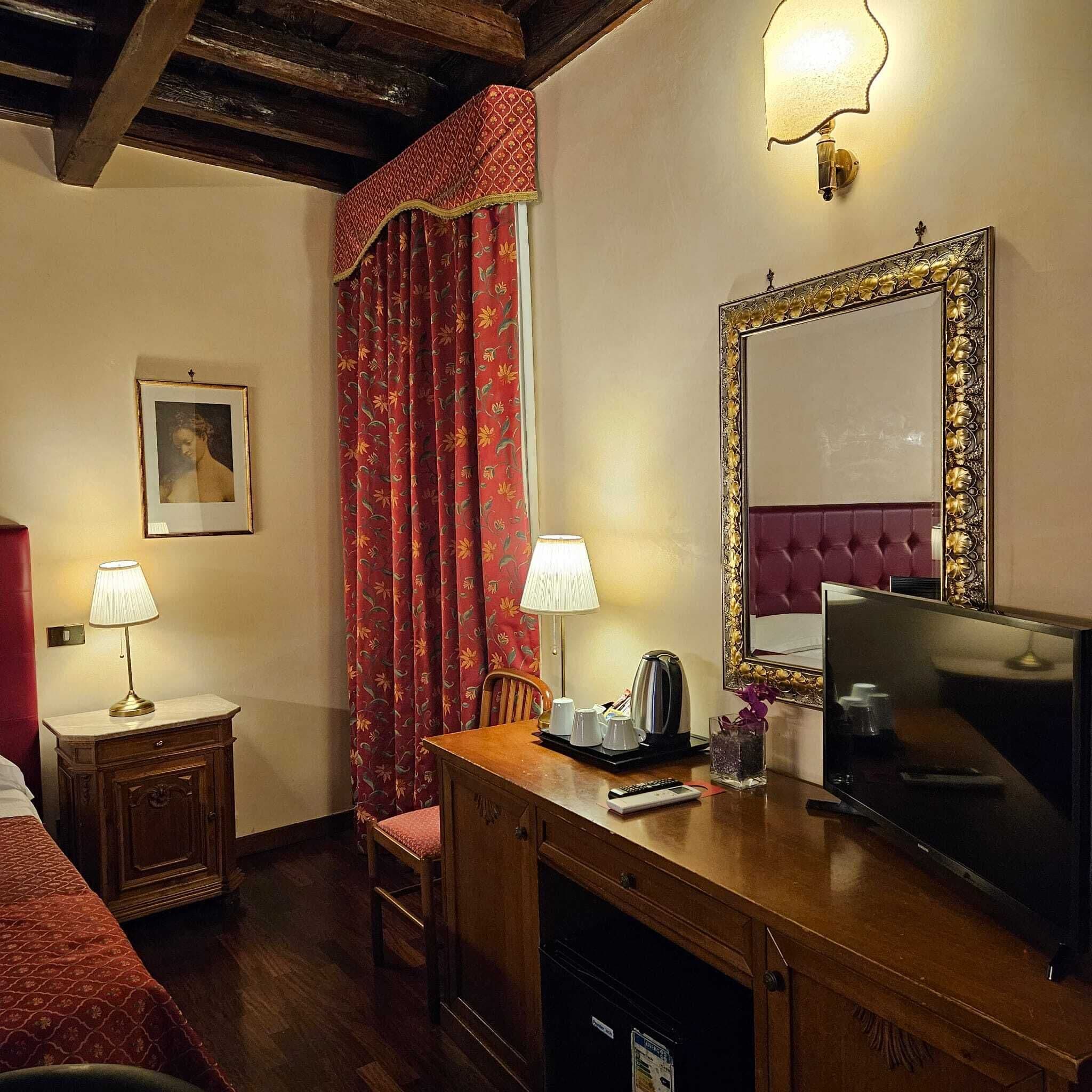 A hotel room with a wooden desk, two bedside lamps, a television, a mirror, and a red floral curtain.