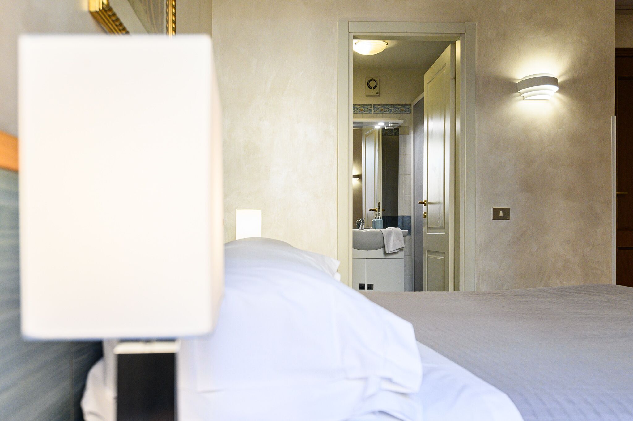 Habitación de hotel con cama, mesita de noche, espejo y baño visible a través de una puerta abierta.