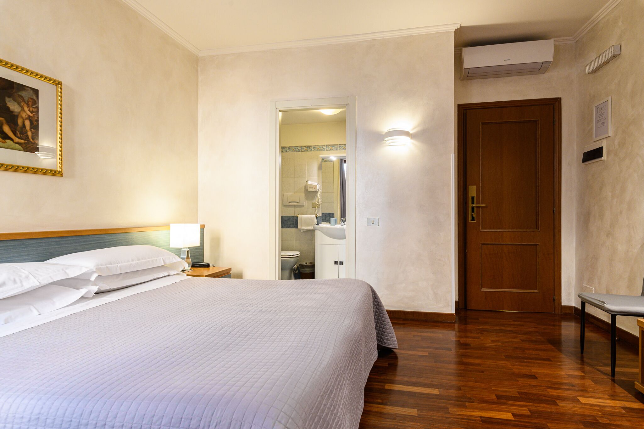 A hotel room with a bed, a nightstand, a bedside lamp, a wooden floor, a door, and a bathroom visible through an open door.