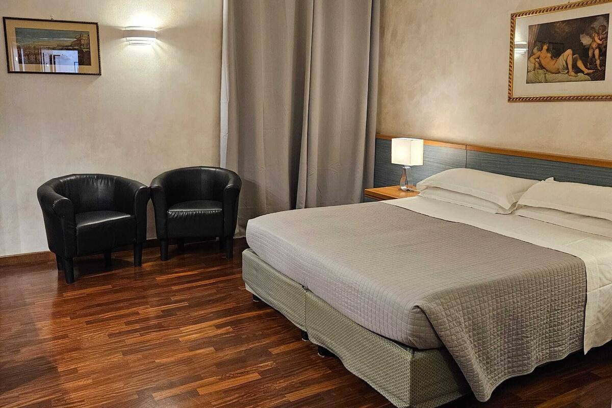 A hotel room with a bed, two armchairs, a painting, and a curtain.