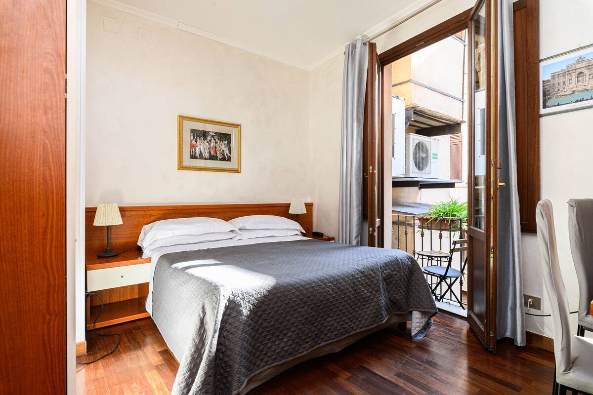 A bedroom with a bed, a nightstand, a lamp, a chair, and a balcony with a view of the outside.