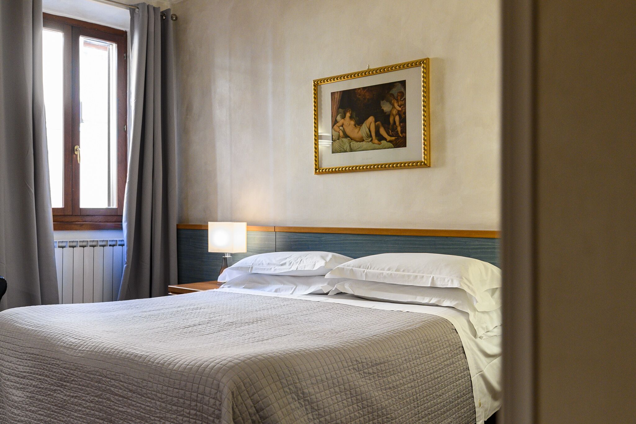 A hotel room with a large bed, a painting on the wall, a window with curtains, and a bedside lamp.