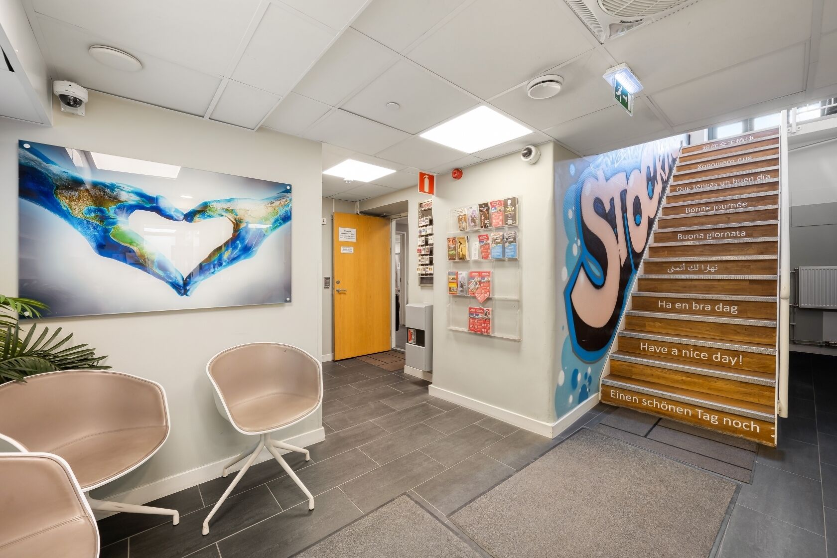 A reception area with a large abstract painting, a wall-mounted display with multilingual greetings, and a staircase with a similar design.