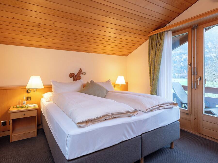 Una cama bien hecha con sábanas y almohadas blancas, dos lámparas de noche, un cabecero de madera, una mesita de noche de madera con una botella y vasos, y vista a un paisaje nevado a través de una puerta de cristal.
