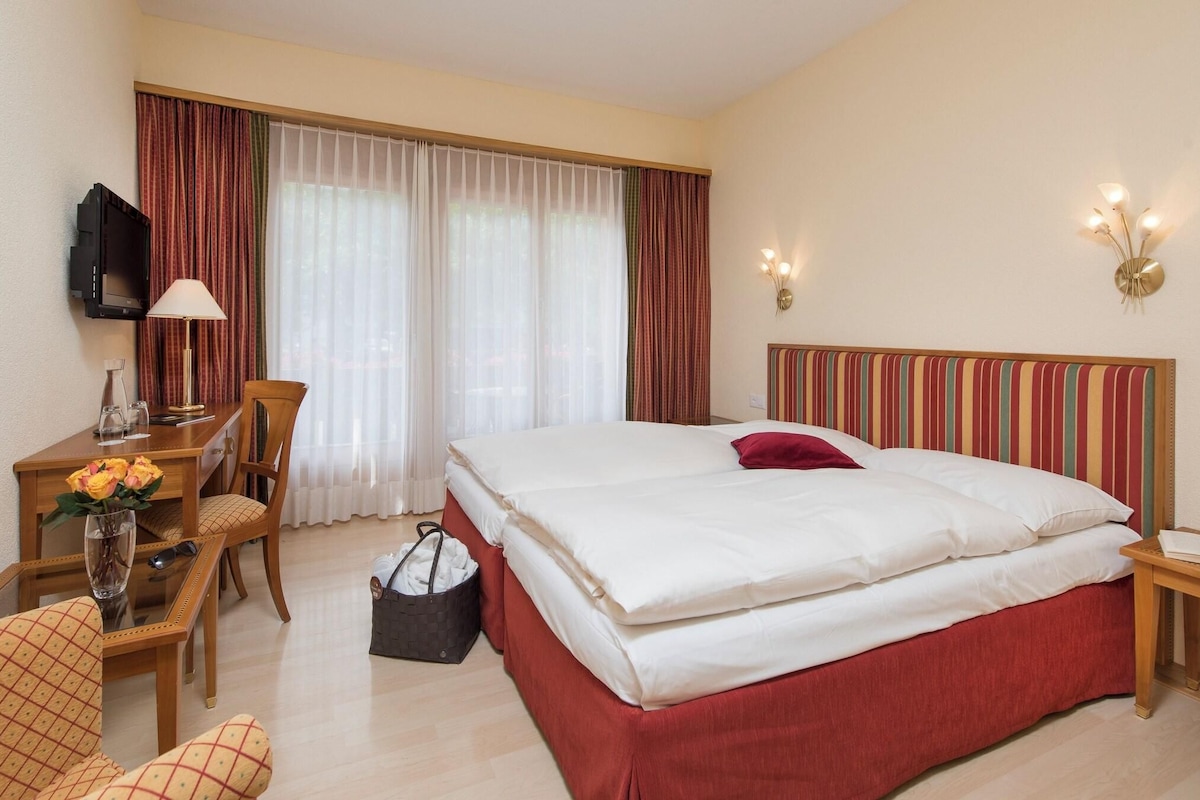A hotel room with a bed, a desk with a lamp, a chair, a TV, and a window with curtains.