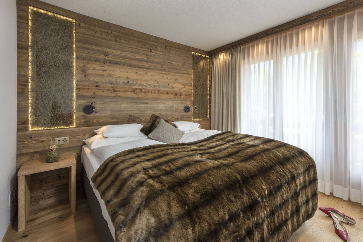 A modern bedroom with a wooden headboard, a large bed with a fur throw, a nightstand with a lamp, and sheer curtains on the window.