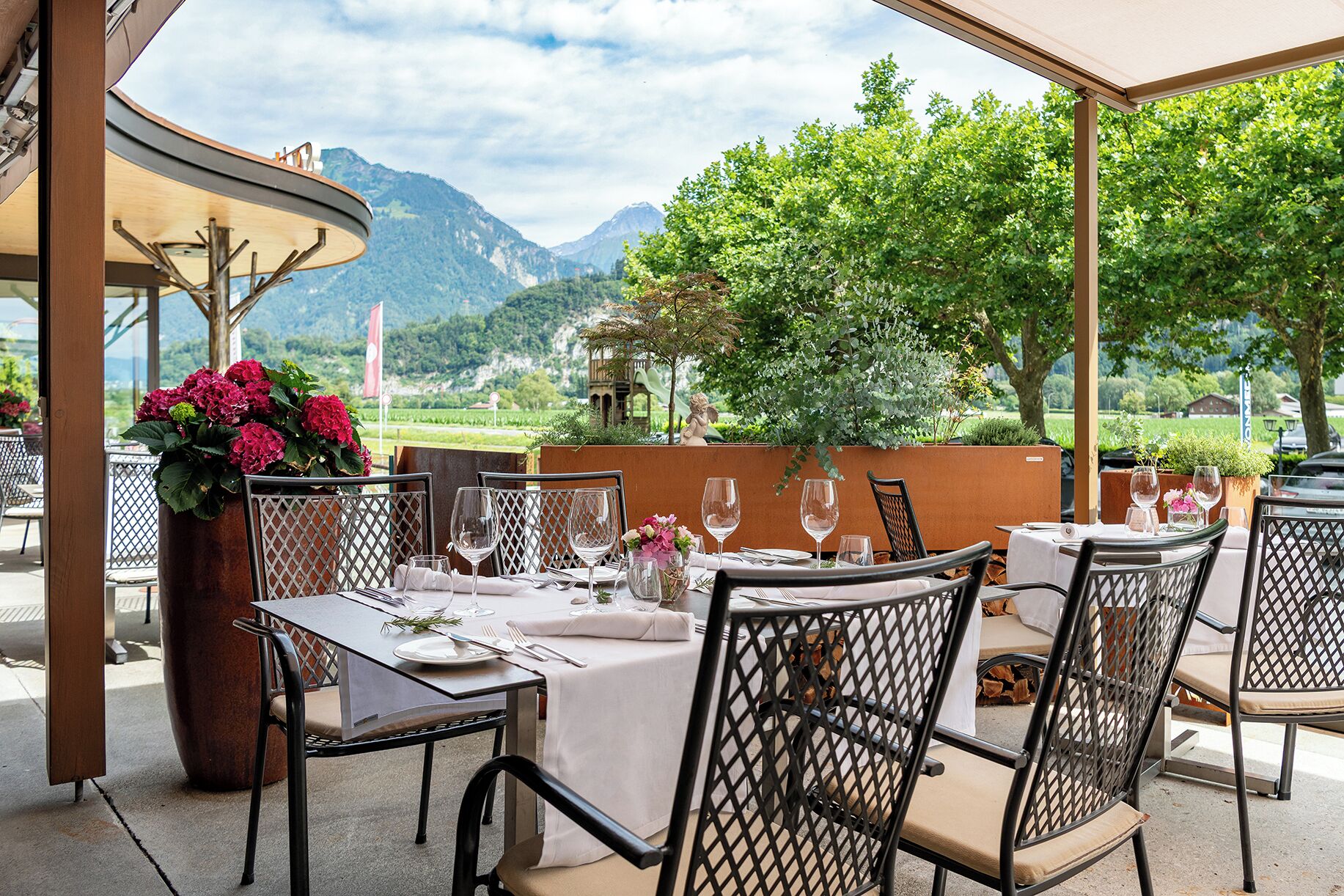 Un espace extérieur aménagé pour les repas, avec des tables pour deux personnes, des chaises et une grande plante en pot ornée de fleurs.