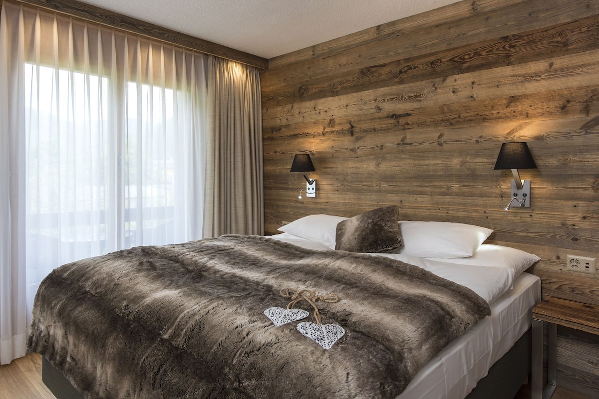 A bedroom with a wooden wall, two bedside lamps, a bed with a dark blanket, and a window with sheer curtains.