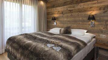 A bedroom with a wooden wall, two bedside lamps, a bed with a dark blanket, and a window with sheer curtains.
