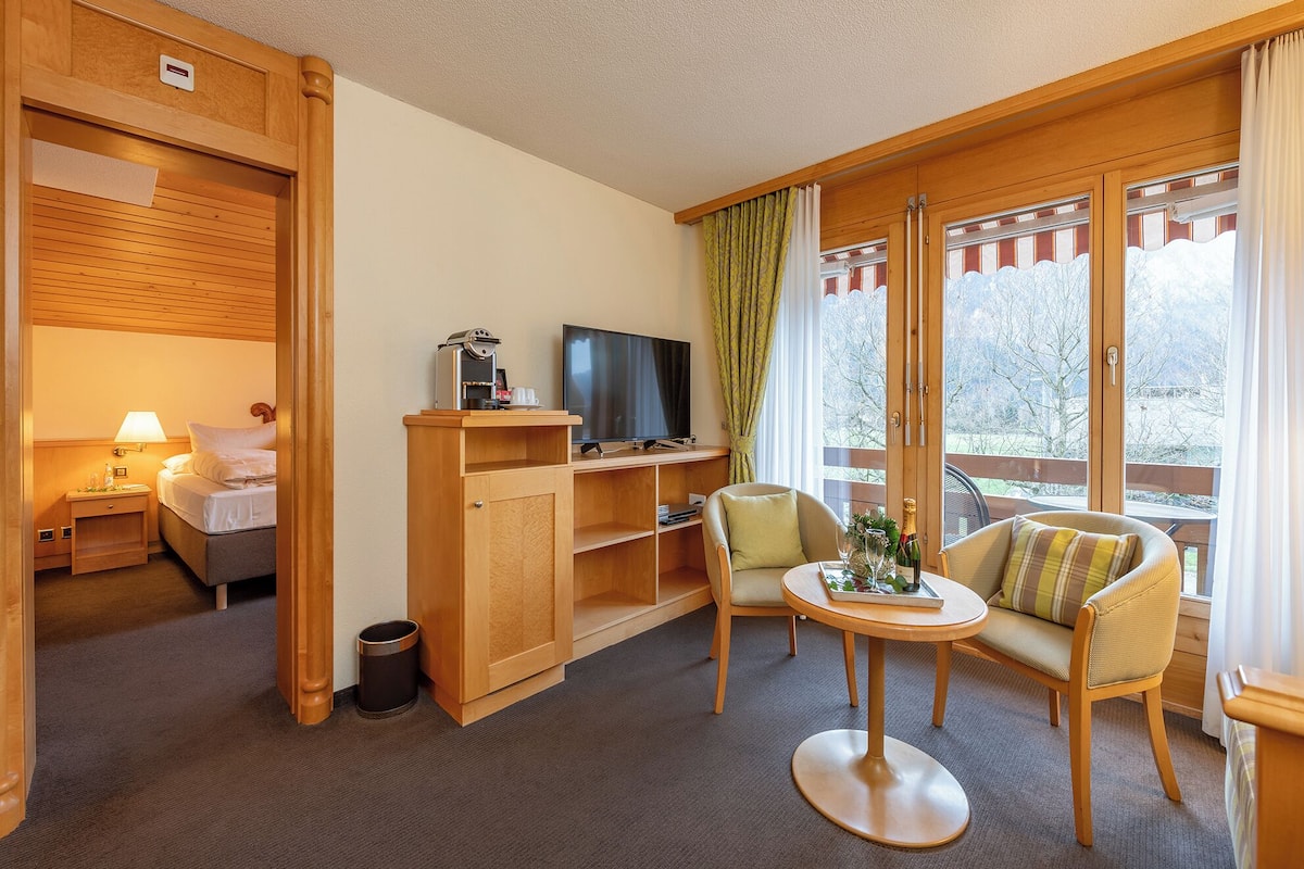 A hotel room with a bed, a TV, a coffee machine, a small table with a bottle, and a window with curtains.