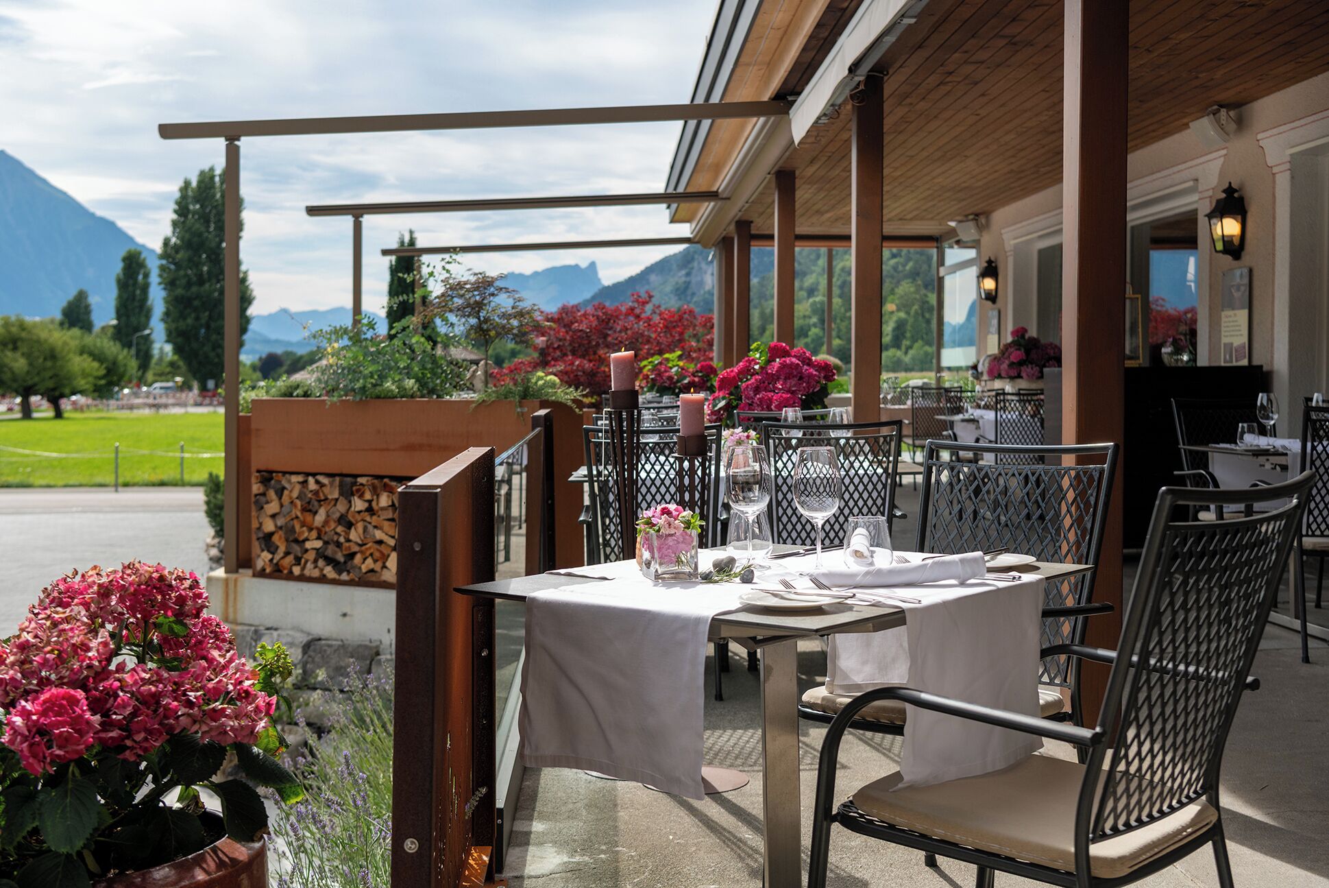 Ein Essbereich im Freien mit Tischen für zwei Personen, umgeben von einer Holzpergola, mit Blick auf die Berge im Hintergrund.