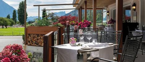 Ein Essbereich im Freien mit Tischen für zwei Personen, umgeben von einer Holzpergola, mit Blick auf die Berge im Hintergrund.