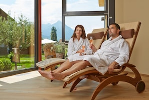 Two people in white bathrobes sitting on a wooden recliner with a glass of wine, in a room with large windows overlooking a garden and mountains. - SALZANO Hotel - Spa - Restaurant (Unterseen)