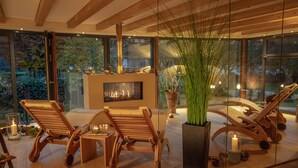 Un intérieur moderne avec une cheminée, des fauteuils en bois et de grandes fenêtres donnant sur l’extérieur.