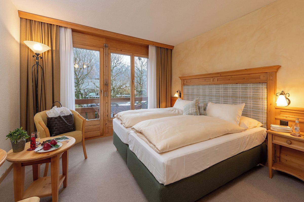 A hotel room with two beds, a wooden headboard, a small table with a chair, a lamp, and a view of the outdoors.