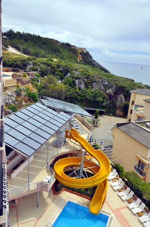 A hotel with a pool, a yellow water slide, and a view of the sea and hills.